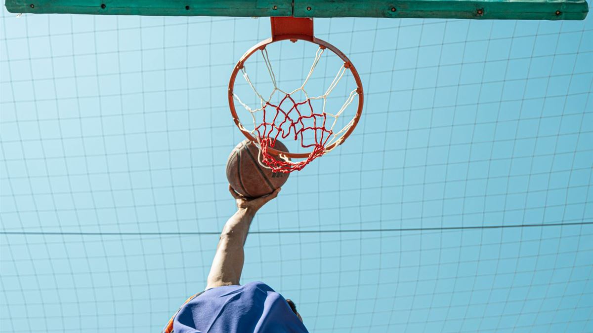 Basketball player seen from below reaching for the rim with the ball on a sunny outdoor court